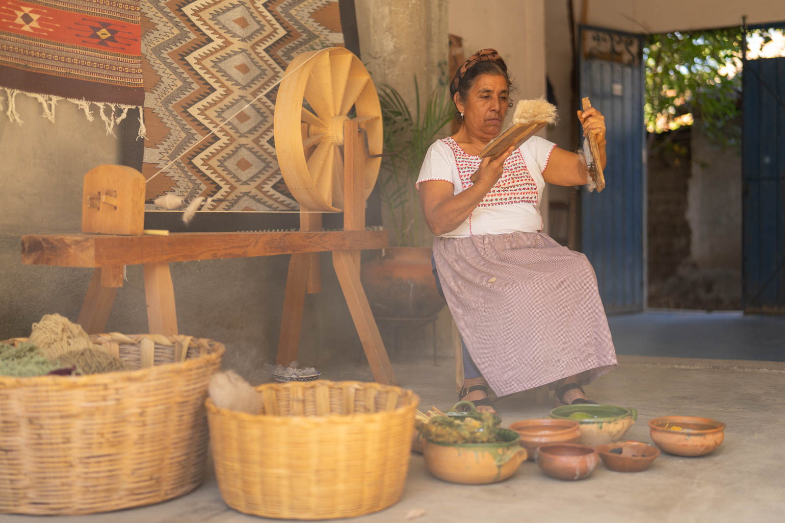 Zapotec artisan carding wool beside a spinning wheel and handwoven rugs in Teotitlán del Valle, Oaxaca